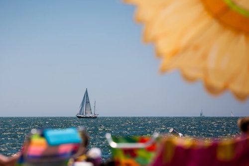 A sailboat crosses the horizon beyond a sun-warmed beach umbrella, seen from the sand.